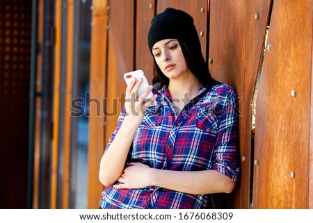 Image of beautiful stylish woman holding mobile phone. Young european girl standing at the street and using cellphone. Woman listening to music with the phone and having fun. Mobile internet concept.