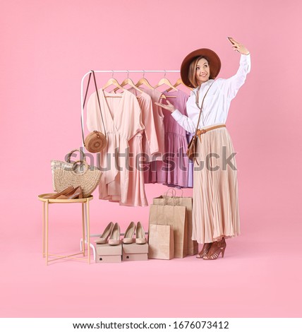 Female stylist near rack with modern clothes recording video on color background