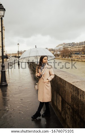 Rain in a winter day in city, woman with transparent umbrella standing on a street near river Seine. Rainy weather, wet roads. Woman with umbrella walking in the rain in Paris. 