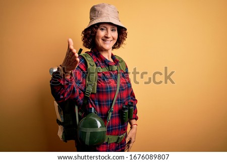 Middle age curly hair hiker woman hiking wearing backpack and water canteen using binoculars smiling friendly offering handshake as greeting and welcoming. Successful business. Royalty-Free Stock Photo #1676089807