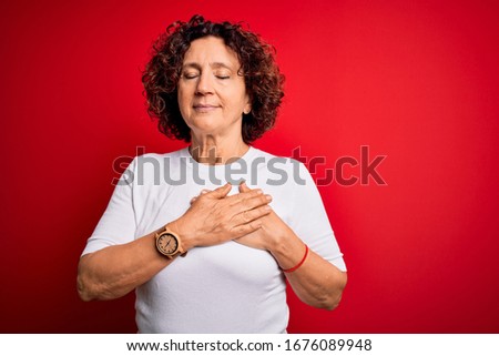 Middle age beautiful curly hair woman wearing casual t-shirt over isolated red background smiling with hands on chest with closed eyes and grateful gesture on face. Health concept.