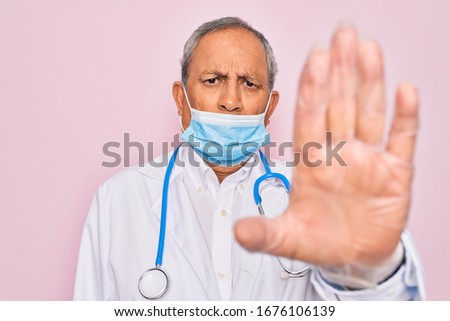 Senior hoary doctor man wearing medical mask and stethoscope over pink background with open hand doing stop sign with serious and confident expression, defense gesture