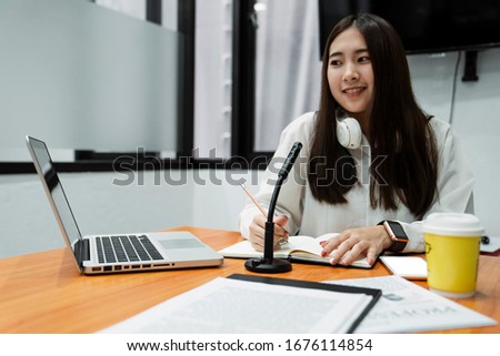 Microphone and laptop on the wooden table in front of smiling woman podcaster in white shirt.