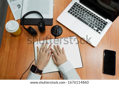 Cropped image of wooden table with black headphone, yellow coffe cup, laptop, notebook, documents, smartphone and microphone in front of the woman in white shirt.