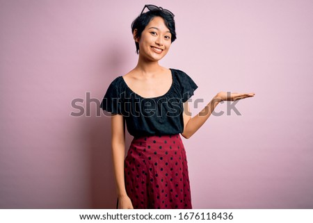 Young beautiful asian girl wearing casual dress standing over isolated pink background smiling cheerful presenting and pointing with palm of hand looking at the camera.