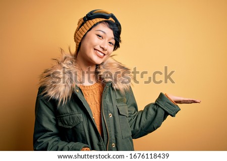 Beautiful asian skier girl wearing snow sportswear using ski goggles over yellow background smiling cheerful presenting and pointing with palm of hand looking at the camera.