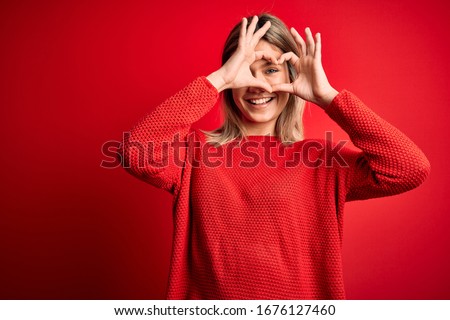 Young beautiful blonde woman wearing casual sweater over red isolated background Doing heart shape with hand and fingers smiling looking through sign