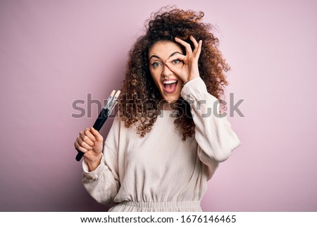 Young beautiful woman with curly hair and piercing applying cosmetic using paint brushes with happy face smiling doing ok sign with hand on eye looking through fingers