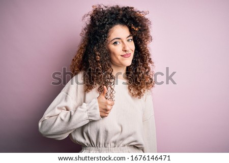 Beautiful woman with curly hair and piercing wearing casual sweater over pink background doing happy thumbs up gesture with hand. Approving expression looking at the camera showing success.