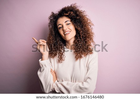 Beautiful woman with curly hair and piercing wearing casual sweater over pink background with a big smile on face, pointing with hand and finger to the side looking at the camera.