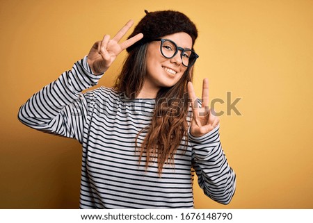 Young beautiful brunette woman wearing french beret and glasses over yellow background smiling looking to the camera showing fingers doing victory sign. Number two.