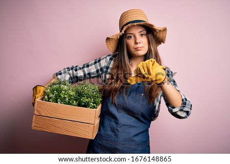Young beautiful brunette farmer woman wearing apron and hat holding box with plants with angry face, negative sign showing dislike with thumbs down, rejection concept