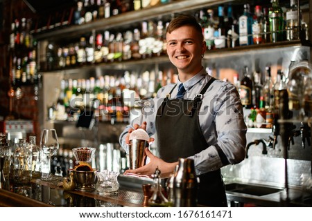 young smiling bartender in black apron preparing to make cocktail. Shelves with bottles of alcohol in background. Royalty-Free Stock Photo #1676161471