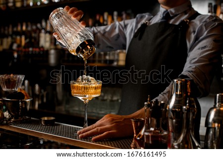 Professional bartender in black apron pours brown alcoholic drink from glassy shaker into glass with ice. Shelves with bottles of alcohol in background. Royalty-Free Stock Photo #1676161495