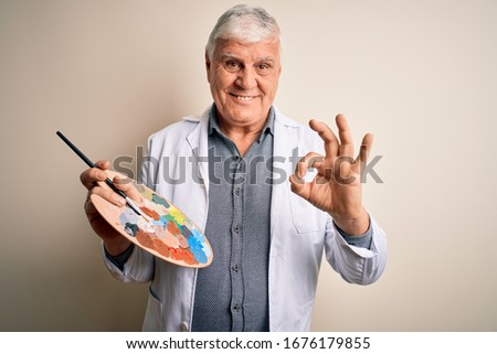 Senior handsome hoary painter man painting using brush and palette with colors doing ok sign with fingers, excellent symbol