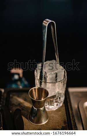 Empty metal jigger and rocks glass with cube of ice. Dark background. Smooth image with shallow depth of field.