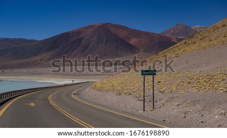 Desert road in the Chilean plateau touching a blue lagoon and leaving behind a high mountain