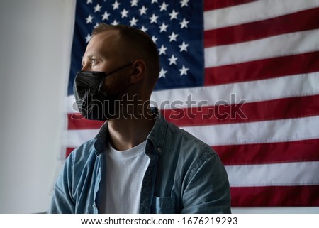 A man in a black medical mask sits on a home quarantine against the backdrop of the American flag. Self isolation due to coronavirus. Measures to curb the spread of Covid-19. Health of the country