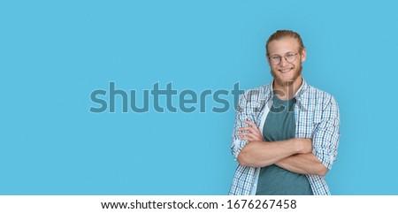 Smiling confident young bearded handsome man hipster wear shirt glasses looking at camera, millennial handsome casual guy student arms crossed isolated on blue studio background, portrait, copy space.