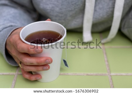 The girl holds in her hand a disposable white cup with black tea. Tea bag inside. Popular takeaway drinks concept. A cup on a green table. Horizontal view.
