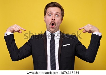 Positive young man with mustache smiling and pointing at himself with index fingers. Handsome man wearing suit gesturing and looking at camera. Self presentation concept. Royalty-Free Stock Photo #1676313484
