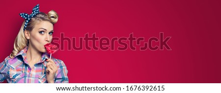 Surprised woman eating heart shape lollipop. Girl in pin up cloth. Blond model at retro fashion and vintage concept. Red color background. Copy space for some text. Wide horizontal banner composition.