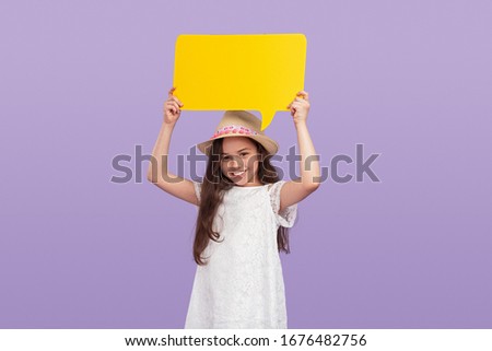 Happy confident preteen girl in white dress and straw hat with brim smiling at camera and holding bright yellow empty banner, while standing alone against light violet background in studio