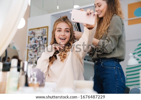 Image of a happy positive young beautiful woman client take a selfie with hair stylist in saloon.