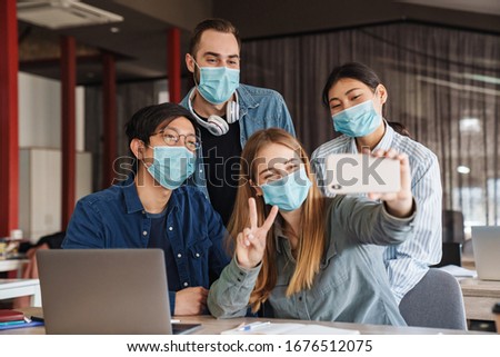 Photo of multinational cheerful students in medical masks taking selfie on cellphone at classroom