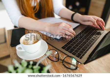 Background. Student girl is sitting at a table in a cafe with a cup of coffee, glasses, a laptop and letters in tasks, typing on a keyboard. Copy space.