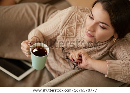Woman in home clothes sitting on the carpet near the sofa using a tablet , drinking coffee from a green cup