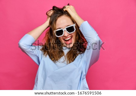 Stylish caucasian girl in a pale blue t-shirt and 8-bit glasses grabs hair and smiles on a pink background. Close up.
