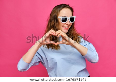Stylish caucasian girl in a pale blue t-shirt and 8-bit glasses and makes a heart with his hands on a pink background. Close up.