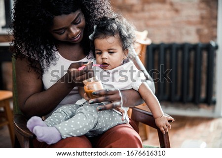 Charming Afro American lady feeding adorable baby girl with spoon stock photo