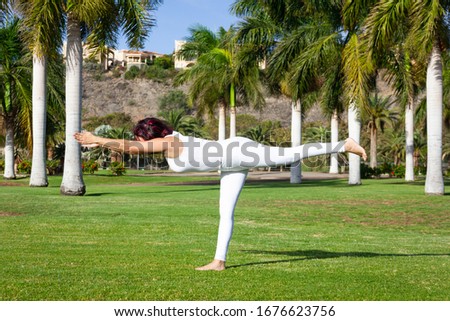 Slim woman in white outfit practices warrior yoga pose on green grass field on sunny day. Female yogi in Virabhadrasana three at park. Balance, equilibrium concepts