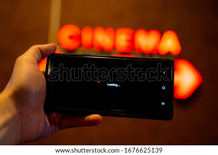 Slow internet, video load and download speed. Watching movie online. Loading icon on screen.Hand with black empty screen mobile phone . Background red wall with cinema signate.