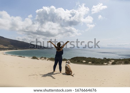 woman raising her arms happy with a large tan and white dog on top of a dune looking at the blue sea, green vegetation, windy and sunny day, landscape
