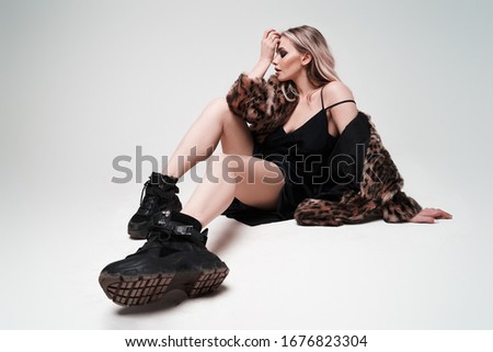 Blonde girl in leopard coat and boots looking at side while sitting in studio