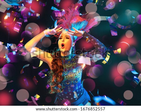 Surprised. Beautiful young woman in carnival, stylish masquerade costume with feathers on black background in neon light, flying confetti. Holidays celebration, dancing, fashion. Festive time, party.