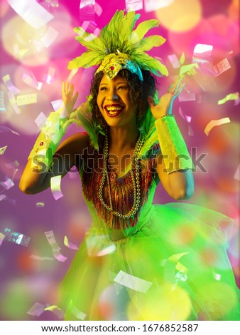 Emotions. Beautiful young woman in carnival, stylish masquerade costume with feathers on gradient background in neon, flying confetti. Holidays celebration, dancing, fashion. Festive time, party.