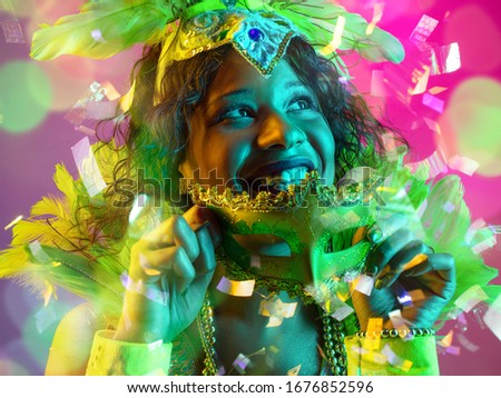 Emotions. Beautiful young woman in carnival, stylish masquerade costume with feathers on gradient background in neon, flying confetti. Holidays celebration, dancing, fashion. Festive time, party.