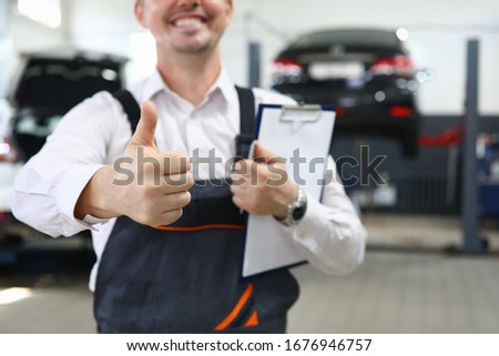 Close-up of mechanic hand showing thumb-up. Happy smile on mans face. Checklist paper. Professional car service station and business concept. Restoration workshop