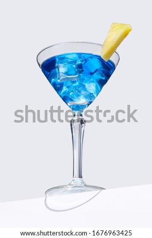 Blue Lagoon cocktail with ice cubes is contained in a martini glass with pineapple slice on the rim and isolated on the table edge. The showy illustrative picture is made on the gray background.