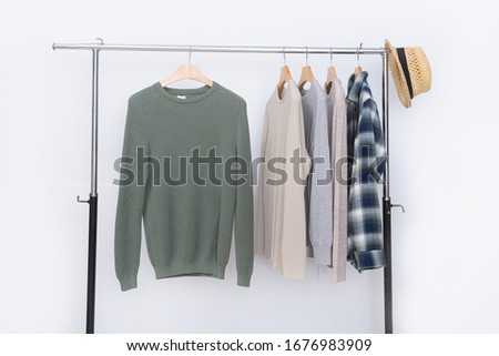 Green sweater with plaid shirt checkered with long sleeved t-shirts hanging and hat on hangers on a white background
