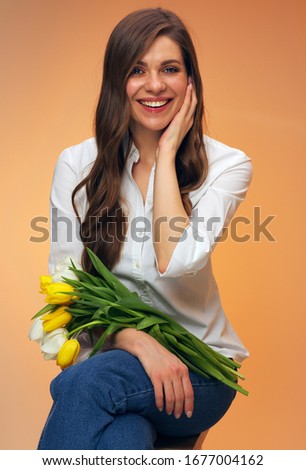 Woman sitting on stool holding tulip flowers and touching her face. Female person isolated portrait on orange background.