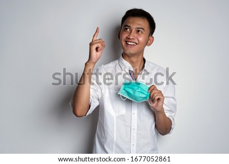 Portrait of friendly asian man showing ecxited expression while holding a health mask with his hand pointing up against white background. Man got a good news expression.  Novel Coronavirus (COVID-19)