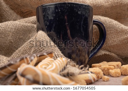 Dark blue ceramic mug of tea , cookies and pieces of cane sugar on a background of coarse fabric. Close up.