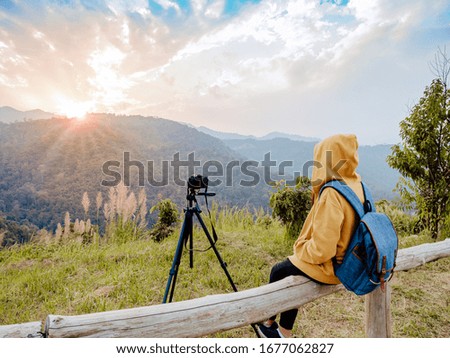 Solo woman with backpack sitting to enjoy mountain view in tropical forest at sunset. And set camera on tripod to take pictures of the atmosphere.