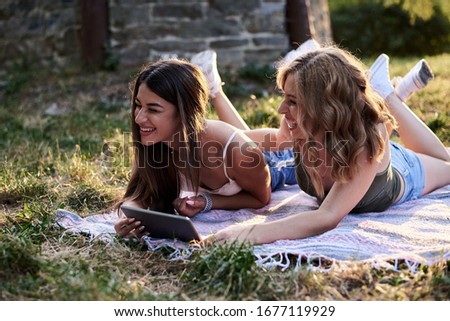 Two sisters, lying on blanket on grass in park in summer, watching fun videos on tablet. Pretty girls, wearing light blue jeans shorts and green beige top, relaxing outside, tickling each other.