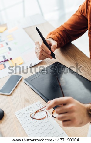 Selective focus of ux designer holding eyeglasses while using graphics tablet near smartphone and layouts on table